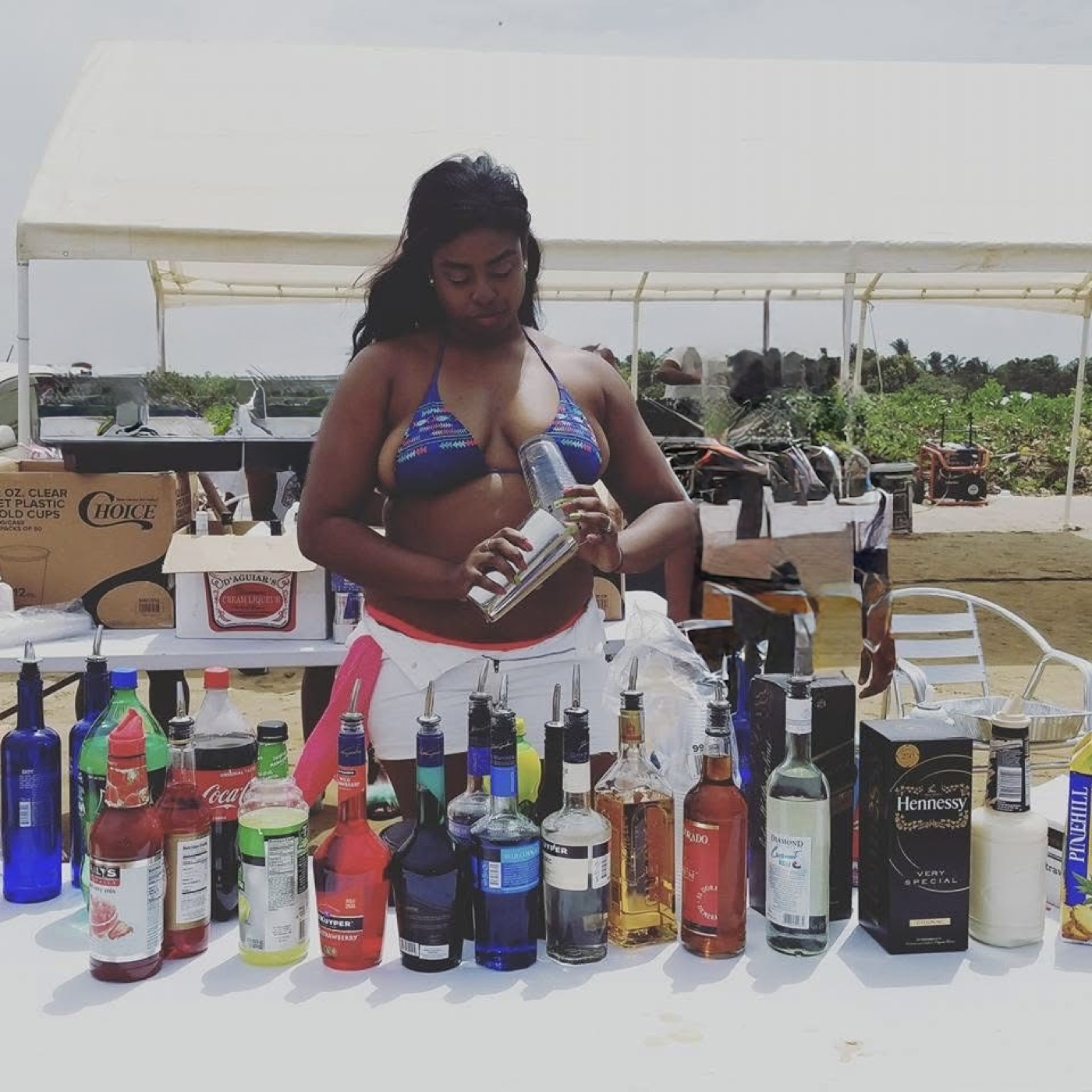 Naomi shaking a cocktail behind a beachside bar in Berbice, bottles lined along the table